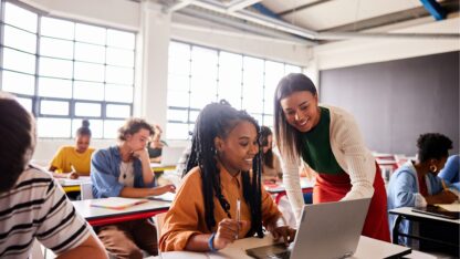 A young woman assisting a student seated inside of a classroom on her laptop.