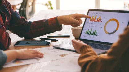 Discussion at a table with fingers pointed at a laptop screen displaying graphs and charts.