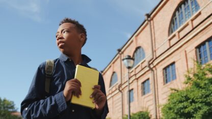 Student looking over his shoulder while walking alongside a building with a book in his hands.