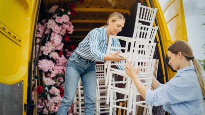 Two event organizers bring chairs, flower arrangements and other party decorations out of the trunk of a van