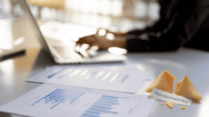 Person working on computer with charts laid out next to them and a fortune cookie that reads "more volume and value" representing predictions for private equity
